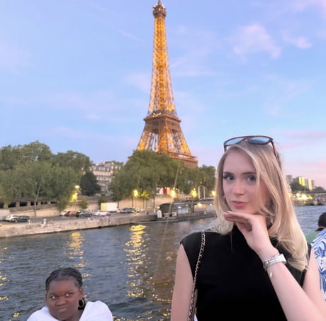 A photo taken at the Eiffel Tower featuring a young woman posing for a selfie in the foreground, while a person in the background is making a funny, expressive face. The juxtaposition of the posed, glamorous foreground and the unintentionally hilarious/dramatic photobomb in the background creates a comedic effect, which the caption 'Hang this in the Louvre' ironically frames as a masterpiece.